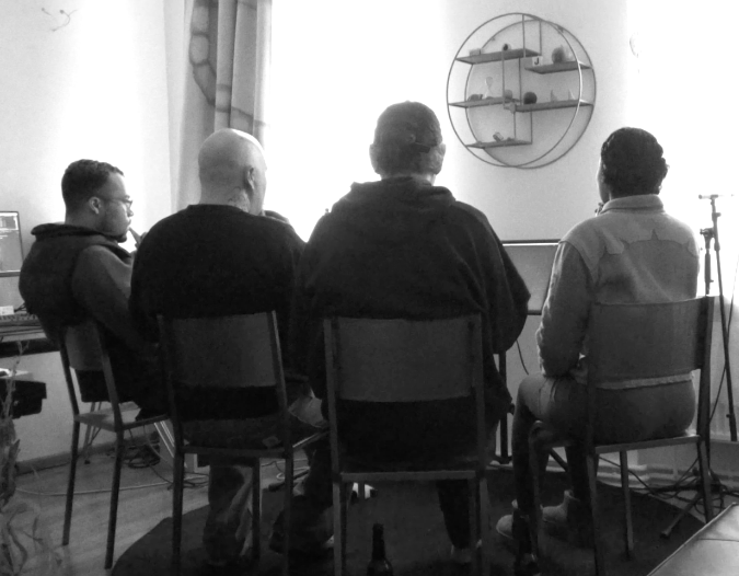 A group of four people are sitting on old fashioned school chairs. They are sitting closely together and are all facing the same direction. They are not sitting in a straight line, but in a sloght curve so that they are clearly working together. They have their back to us so it is unclear what they are doing, but one head is turned in way that we can  see that they are playing a bottle.