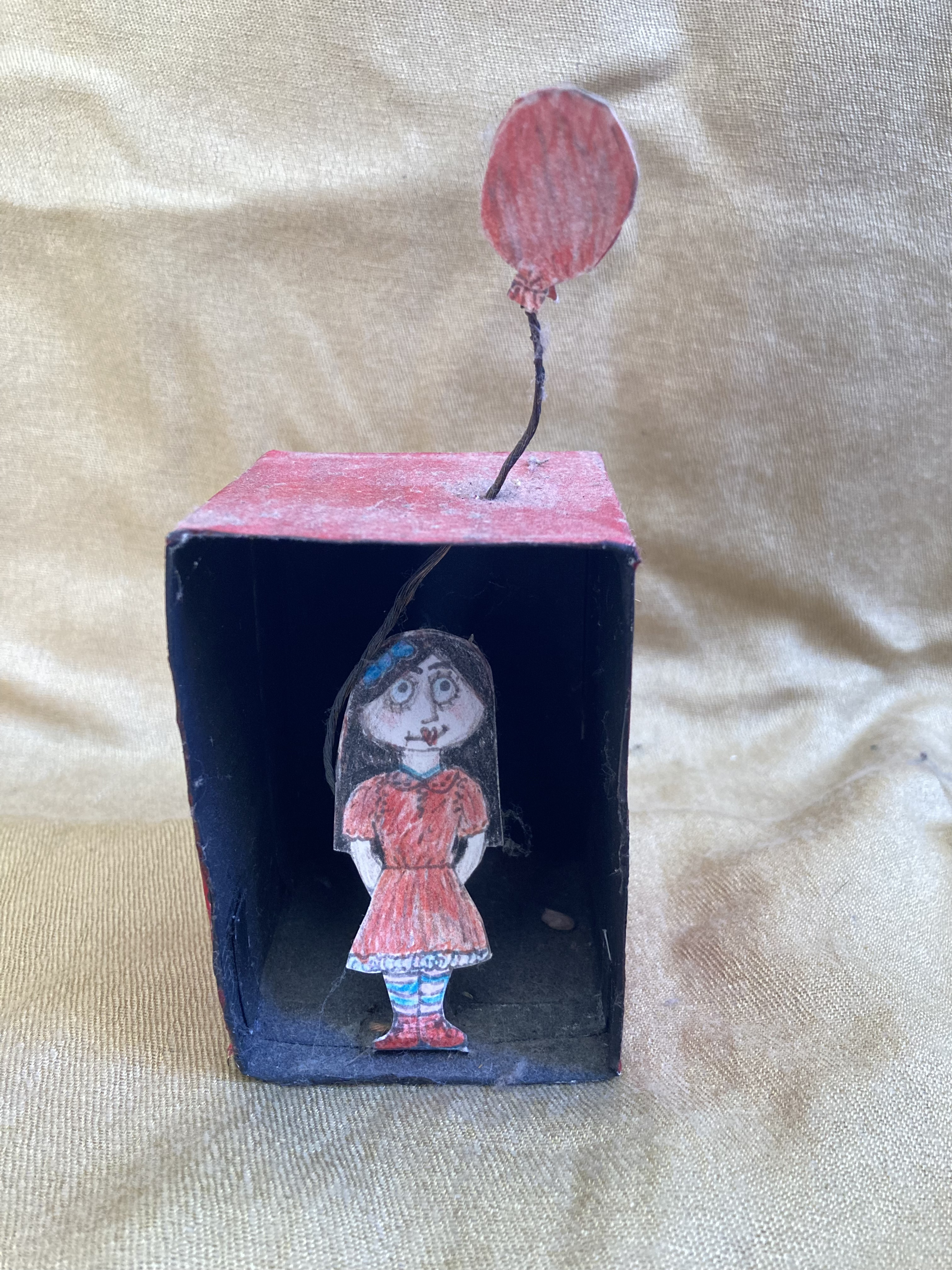 A crayon drawing of a yound girl. Her lips are a love heart and her eyelashes are long. She stands in a lightless box. Out of the top of the box a crayon balloon floats.