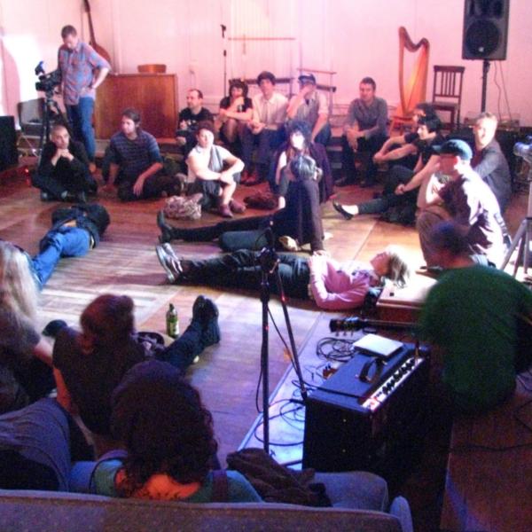 a still from the 'Lines of Flight' documentary. Audience members sit in a circle listening carefully. Some lie with their back on the floor, legs spread into the stage area in the middle of the circle.