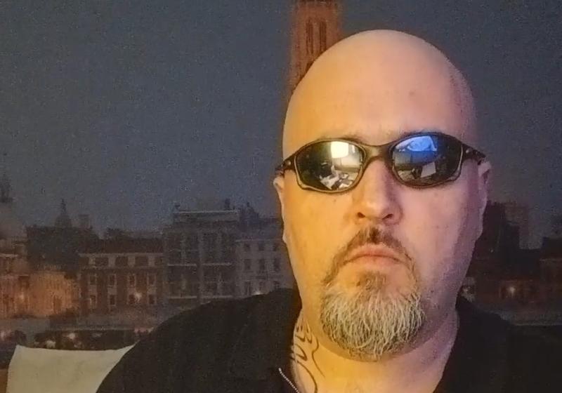 Jeremy is wearing sunglasse at night. They are closely fitting wrap around sunnies. His head is shaved smooth. Behind him is a European square with a large tower of either church or municipal function. His face is serious and stern.