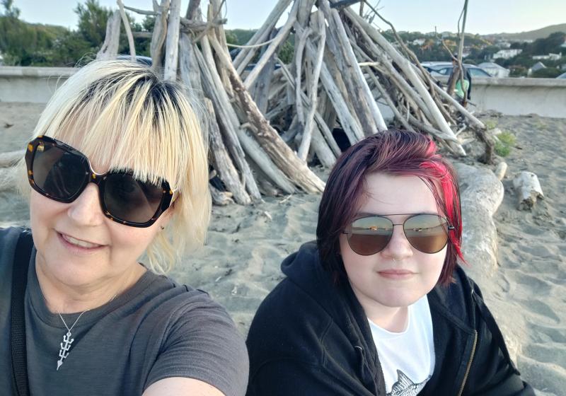 Pip and Bo sit on a sandy beach. They are in front of drift wood stacks arranged for a bonfire. They are relaxed and easy together.