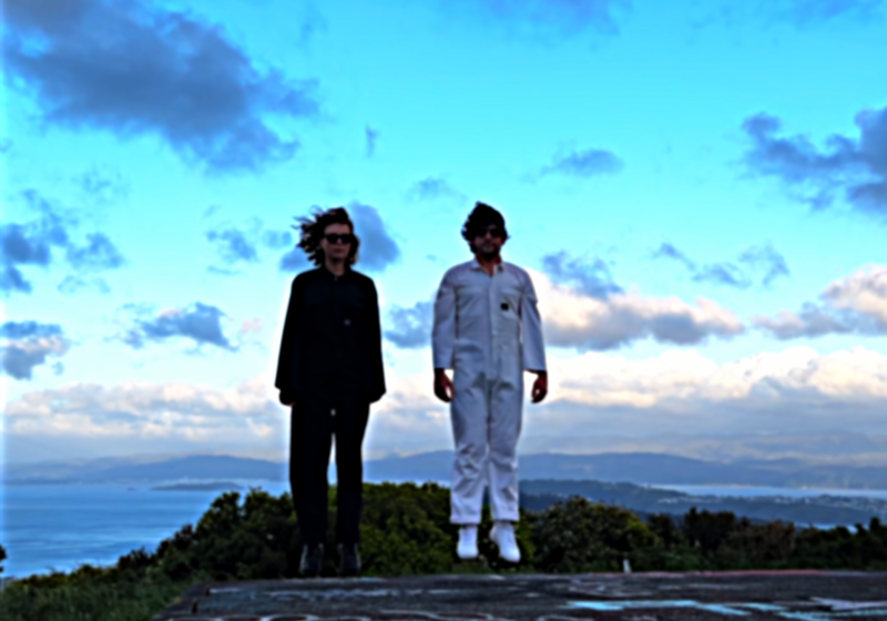 A photo of the duo - it is sunset. They are wearing matching boiler suits, but one is black and the other white. They are jumping in the air and the photo is taken so that it looks like they are levitating.