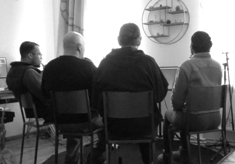 A group of four people are sitting on old fashioned school chairs. They are sitting closely together and are all facing the same direction. They are not sitting in a straight line, but in a sloght curve so that they are clearly working together. They have their back to us so it is unclear what they are doing, but one head is turned in way that we can  see that they are playing a bottle.