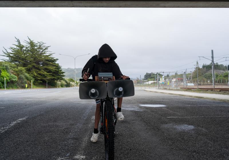 A kid on their bike by the train tracks. The kid is holding a phone and tweaking some knobs on some audio gear mounted on the handle bars. There are two huge siren speakers hanging off the front of the bars.