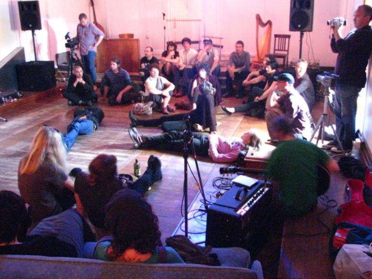 a still from the 'Lines of Flight' documentary. Audience members sit in a circle listening carefully. Some lie with their back on the floor, legs spread into the stage area in the middle of the circle.