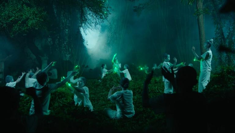 A still from a film by Korakrit, featuring a gathering of 10+ people in a forest touching green lasers/lights beaming