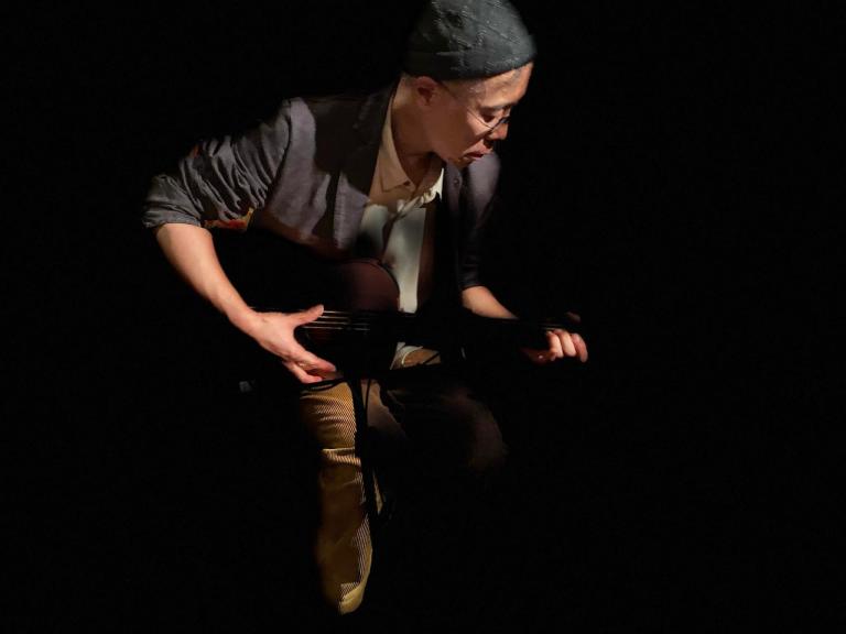 Kawol plays a stringed instrument with a black background