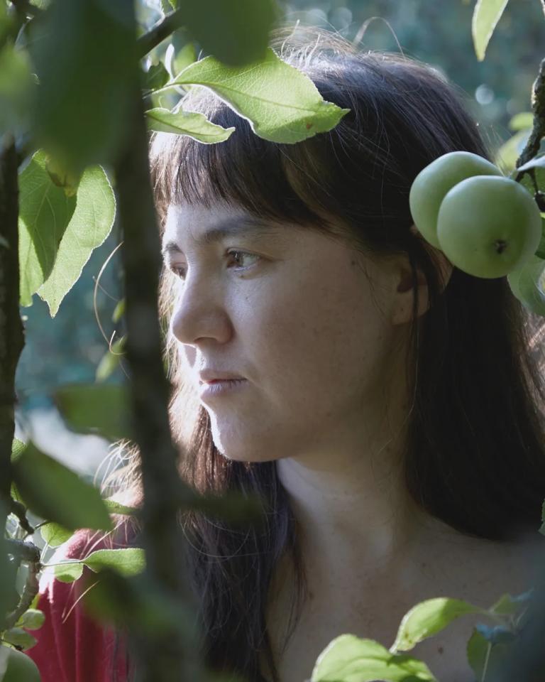 A close up of Delphine Dora amongst greenery looking calmly into the distance
