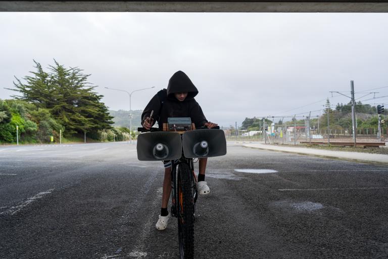 A kid on their bike by the train tracks. The kid is holding a phone and tweaking some knobs on some audio gear mounted on the handle bars. There are two huge siren speakers hanging off the front of the bars.