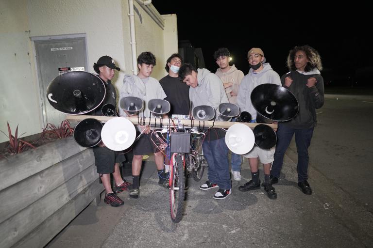 ai-Rhys and friends pre siren battle, Lower Hutt, 2022, a group gather behind a bike. A long wooden plank is stretched out across the front of a bike, supporting 12 speakers of different sizes. The length of assembled sirens rivals the full length of the bike itself, and precariously balances in front the handlebars, threatening to topple the bike.