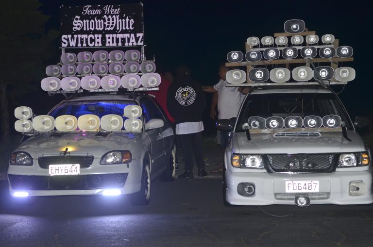 two cars are pictured in battle formation: front-on and side-by-side. Sirens are mounted at the base of the windscreen, in the fender, and in three- and four-tiered stacks on the roof. The car on the left gains even further height with a sign that reads in a mix of cursive, gothic, and all-caps serif font (again, in three tiers) ‘Team West | Snow White | Switch Hittaz’.