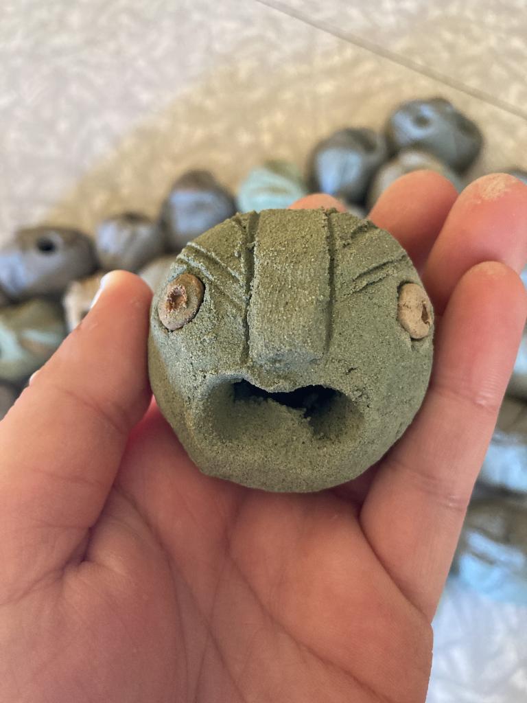 the clay head has round fierce eyes and their open mouth is urgent